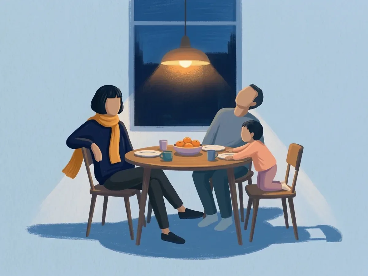 Under a hanging lamp, the same person leans back at a round table beside a partner and a child kneeling on a chair. Oranges, plates, and cups are on it.